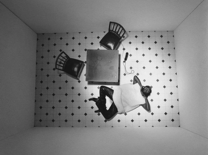 Black and white overhead shot of a woman lying on a tiled floor with chairs and a table in a bright room.