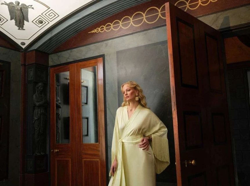 Elegant adult woman in a light yellow belted kimono-style dress stands in an ornate indoor hallway with dark wood and marble accents.