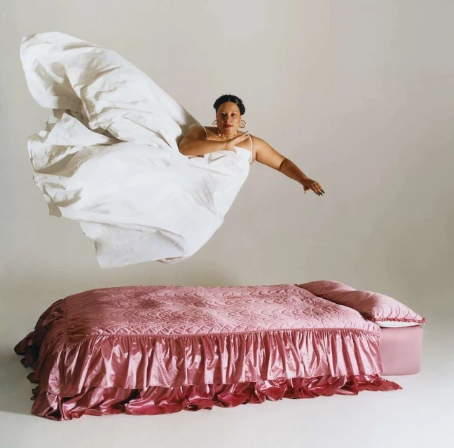 A woman in a white dress floating above a pink bed against a neutral backdrop in a studio-style setup, creating a surreal levitation effect.