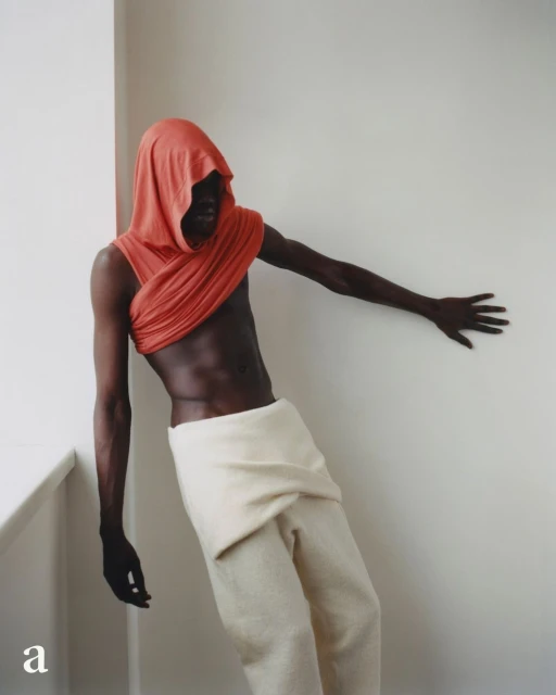 An adult male poses indoors wearing a draped orange hood and neutral-toned wrap pants in a fashion editorial shot in natural lighting.