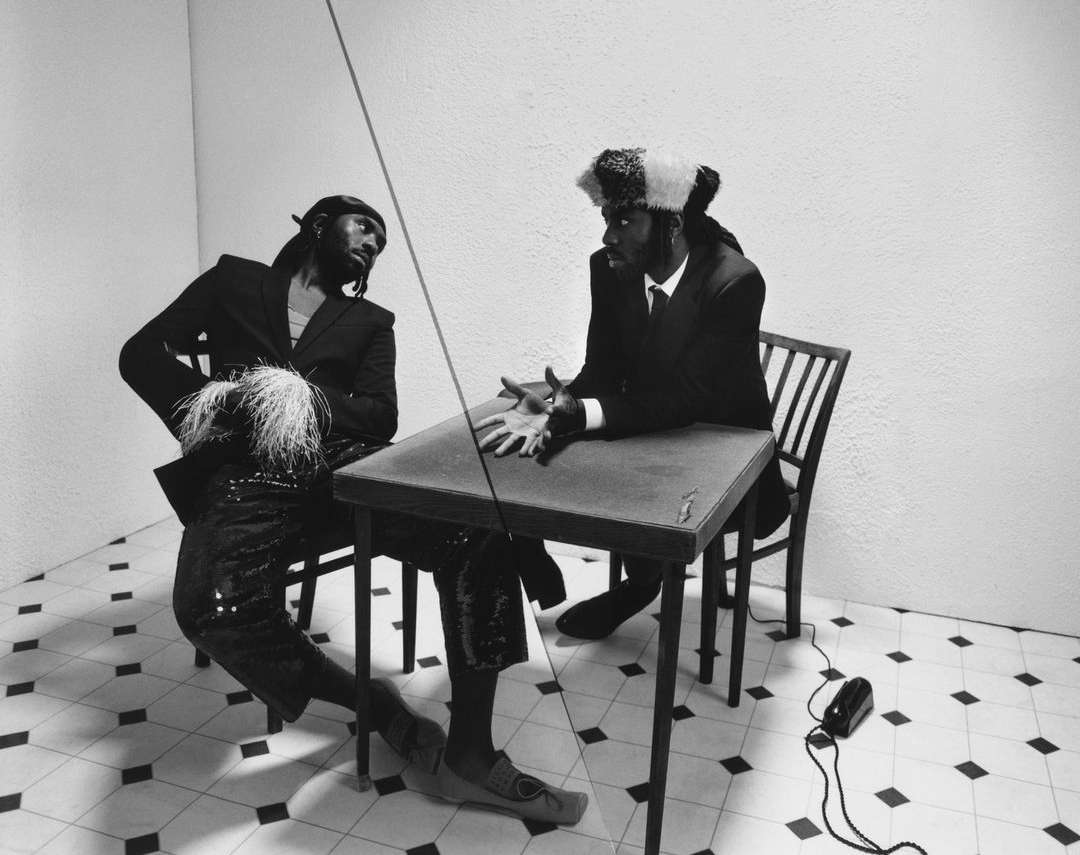 Two men in suits sit at a table in a minimalist setting. One man wears a distinctive hat with white accents. Black and white photo.