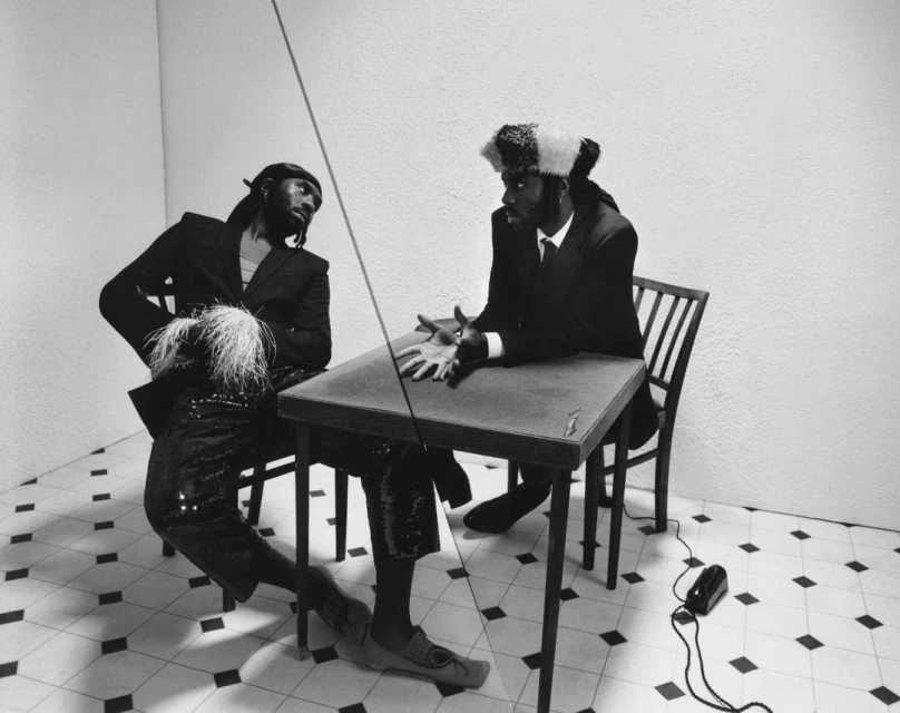 Two men in suits sit at a table in a minimalist setting. One man wears a distinctive hat with white accents. Black and white photo.