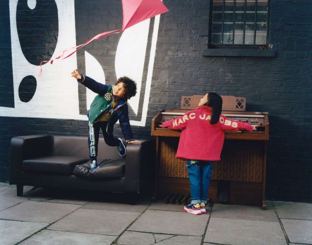 A young child flies a kite near a girl playing an organ outdoors, with a black brick wall and graffiti background in an urban setting.