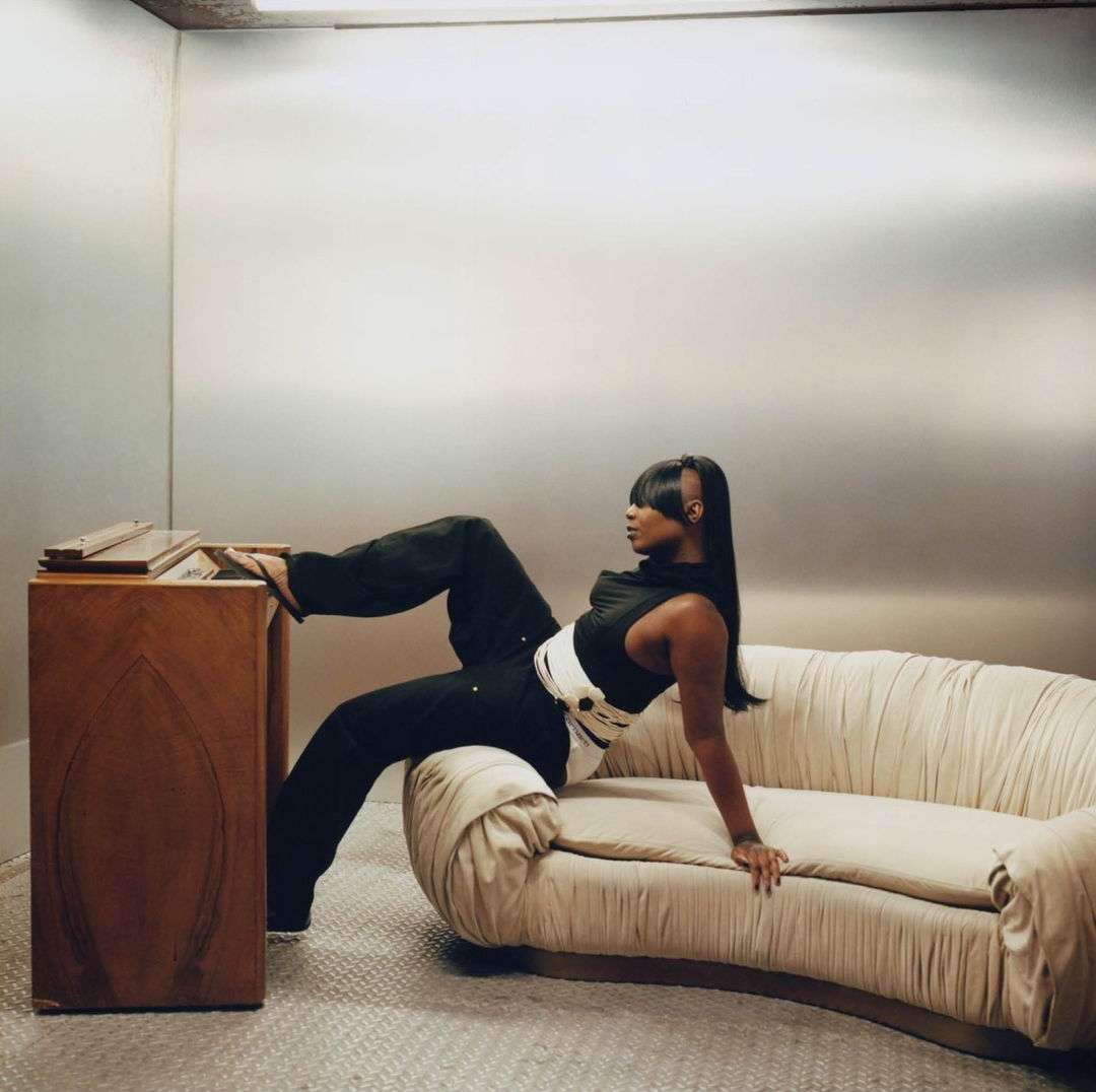 Stylish woman in black outfit poses on cream couch against a metallic background inside an enclosed space with a wooden cabinet.