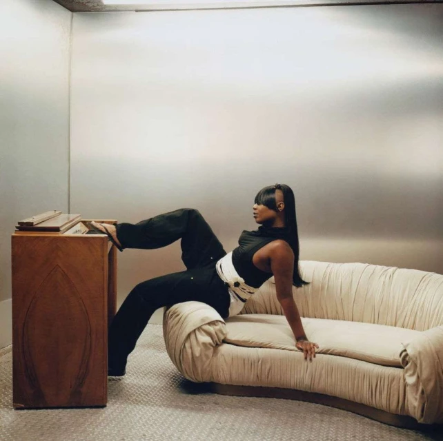 Stylish woman in black outfit poses on cream couch against a metallic background inside an enclosed space with a wooden cabinet.