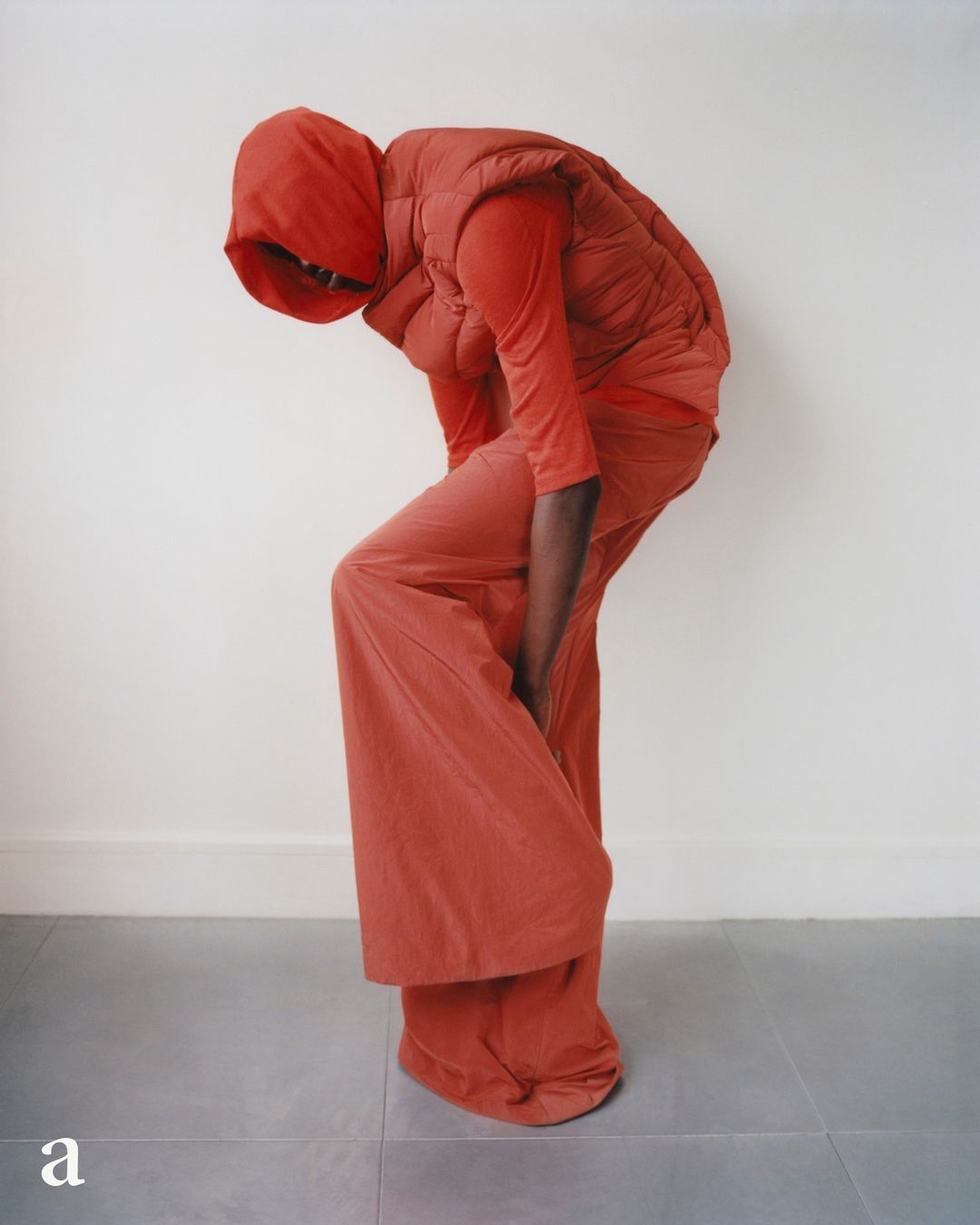 Person in matching red outfit poses in a studio with a white wall and grey tile floor. They are wearing a puffer vest and loose pants.