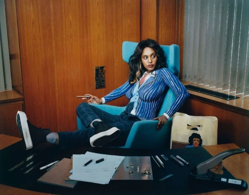 Elegant woman lounges in a turquoise chair in a wood-paneled room wearing a blue pinstripe suit and black platform shoes while smoking a cigar.