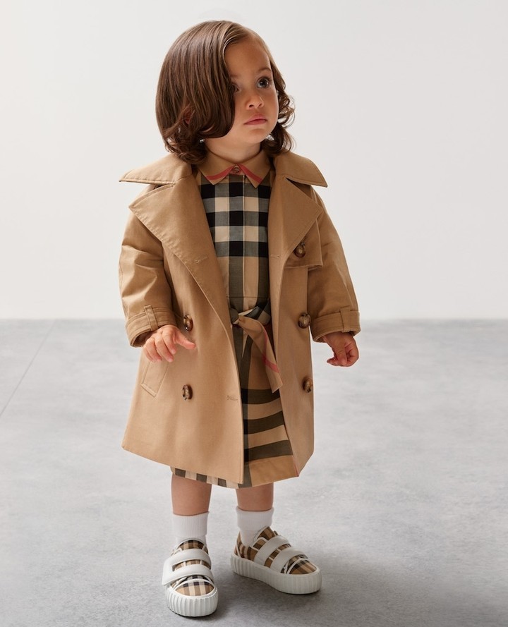 Child in Burberry trench coat and dress, posing in a studio. Classic, timeless style for kids fashion. Neutral backdrop, soft lighting.