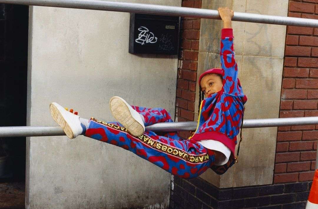 Child in red and blue patterned tracksuit hanging from a metal bar in an urban setting, wearing white sneakers and a red hat.