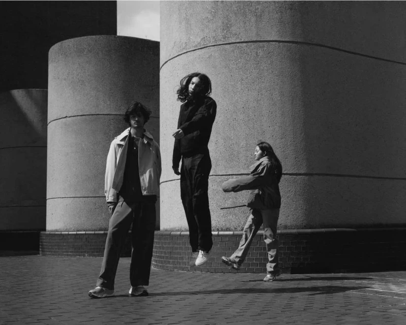 Three people in an urban setting: two on the ground and one levitating between concrete structures, captured in black and white.