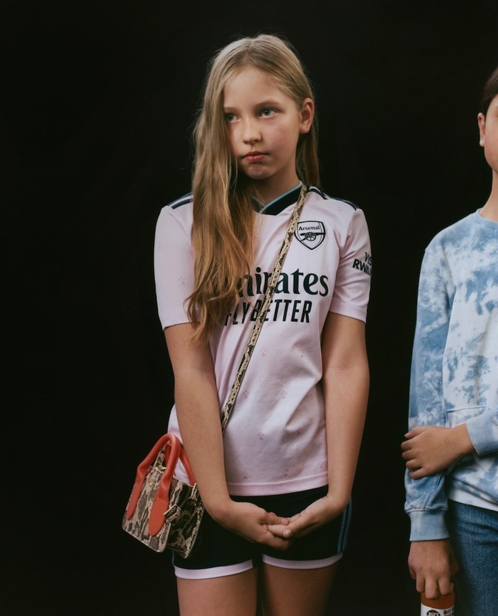 Young person wearing a pink soccer jersey and a small purse stands in front of a black background with another person to the side.