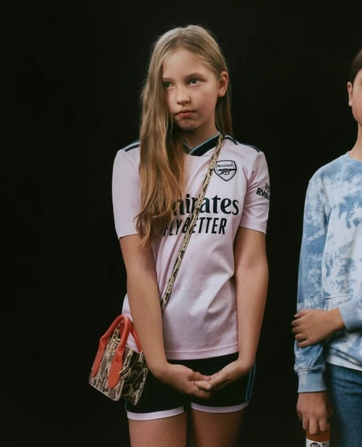 Young person wearing a pink soccer jersey and a small purse stands in front of a black background with another person to the side.