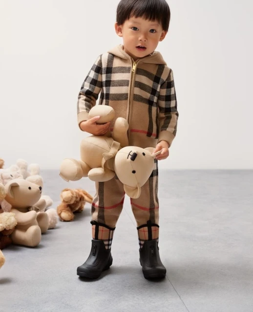 Toddler dressed in plaid onesie holding stuffed animal bear, several teddy bears on floor, child fashion, eye-level angle.