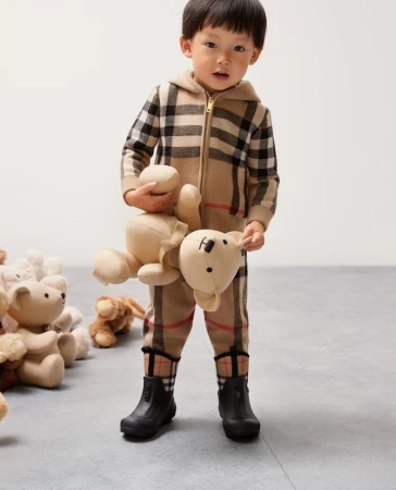 Toddler dressed in plaid onesie holding stuffed animal bear, several teddy bears on floor, child fashion, eye-level angle.