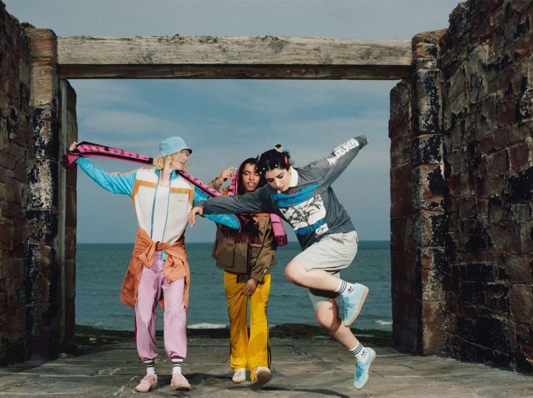 Three adult females pose in bright athletic wear in front of a weathered stone structure and ocean backdrop, creating a casual, sporty editorial.