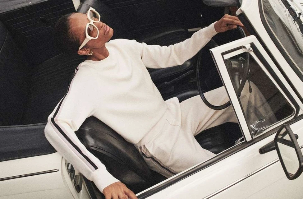 Woman in white sunglasses and a matching tracksuit poses in a vintage white convertible car with black interior on a sunny day.