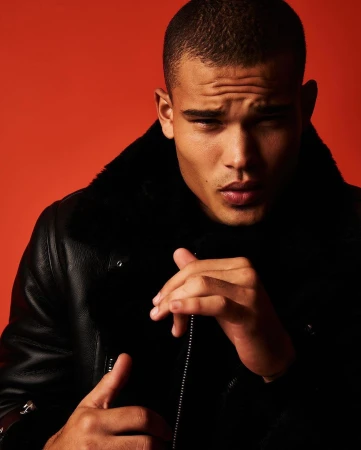 Close-up portrait of a young adult man in a black jacket with fur collar, against an orange background, looking at the camera.