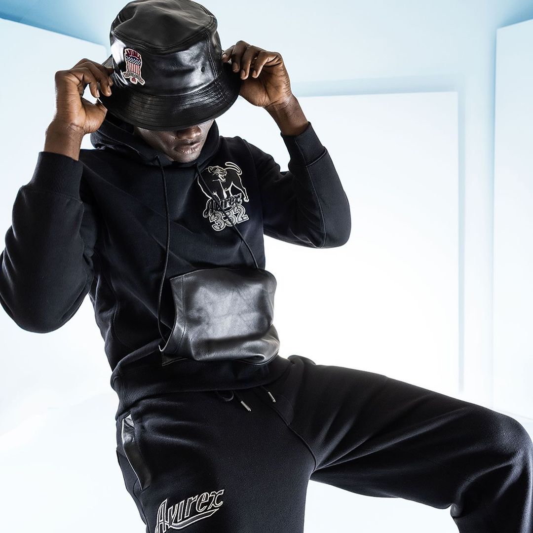 Adult male wearing a black bucket hat, hoodie with graphic, black pants, and a leather crossbody bag against a bright white background.