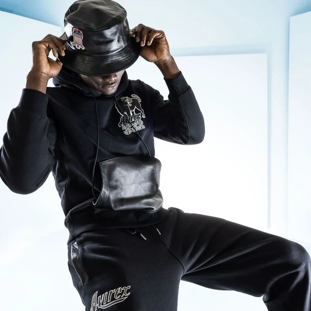 Adult male wearing a black bucket hat, hoodie with graphic, black pants, and a leather crossbody bag against a bright white background.