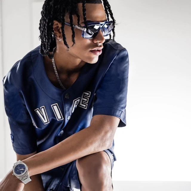 Young adult male poses indoors wearing a blue baseball jersey, shorts, sunglasses, chain necklace, earrings, and a watch with a white background.