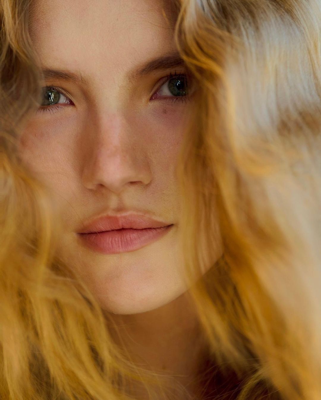 Close-up portrait of a young adult woman with wavy, golden-blonde hair framing her face, and subtle makeup highlighting her features.