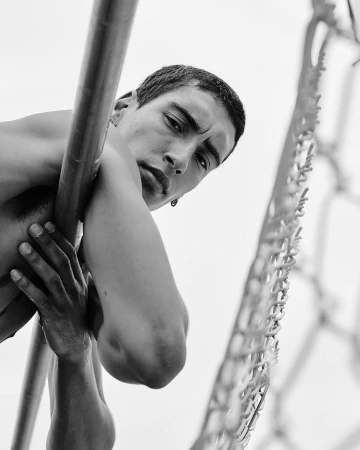 Striking black and white portrait of a young adult male clinging to metal bars, shot from a low angle with a cloudy sky background.
