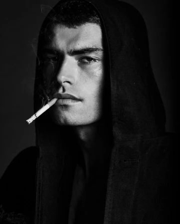 Close-up portrait of a man in a black hooded sweatshirt smoking a cigarette, photographed in monochrome.
