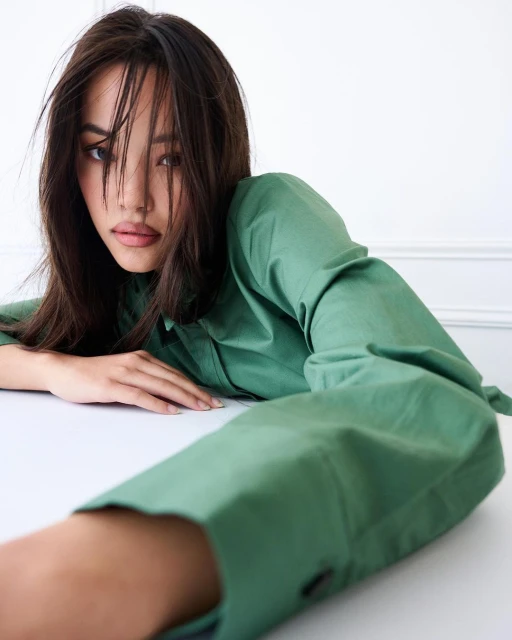 Portrait of a young adult female with dark hair, wearing a green collared shirt and posing against a plain white wall and table.