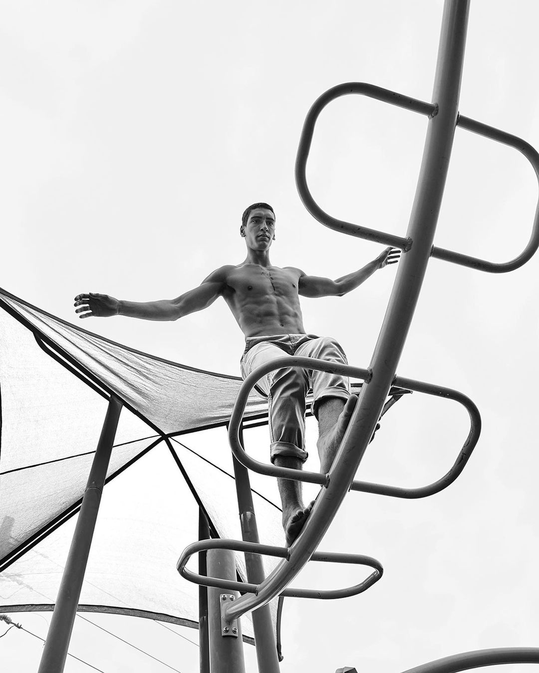 Black and white portrait of a muscular adult male balancing on metal playground equipment with arms outstretched against a cloudy sky.