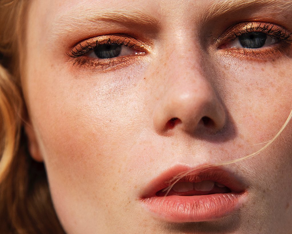 Close-up of a young adult female with fair skin, freckles, and gold eyeshadow, showcasing a beauty and fashion aesthetic.