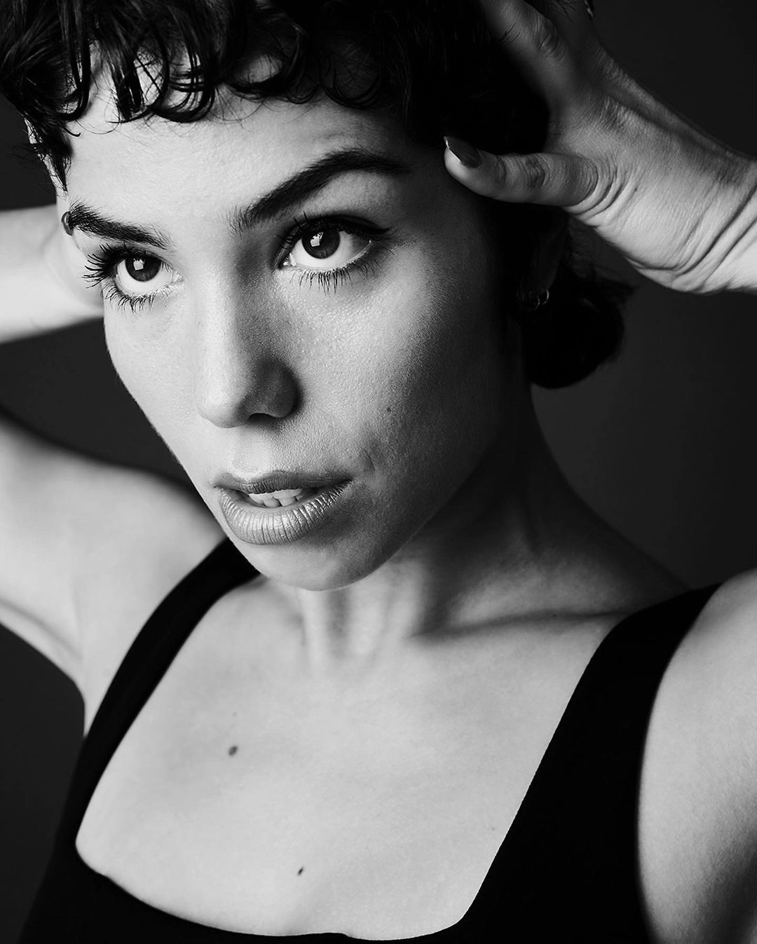 Close-up black and white portrait of a woman with short dark curly hair holding her hands to her head.