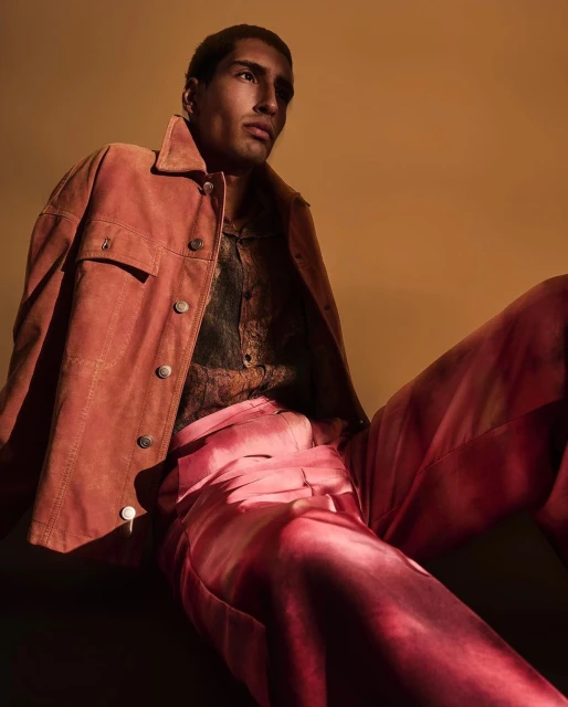 Portrait of an adult male model wearing a rust jacket, printed shirt, and pink trousers, posing in a studio with a tan background.