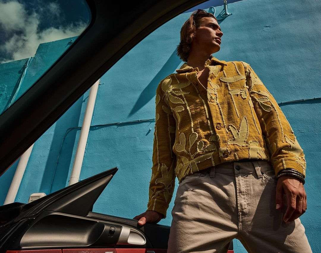 Stylish man in an ochre floral jacket posing against a bright blue wall next to an automobile under the afternoon sun.