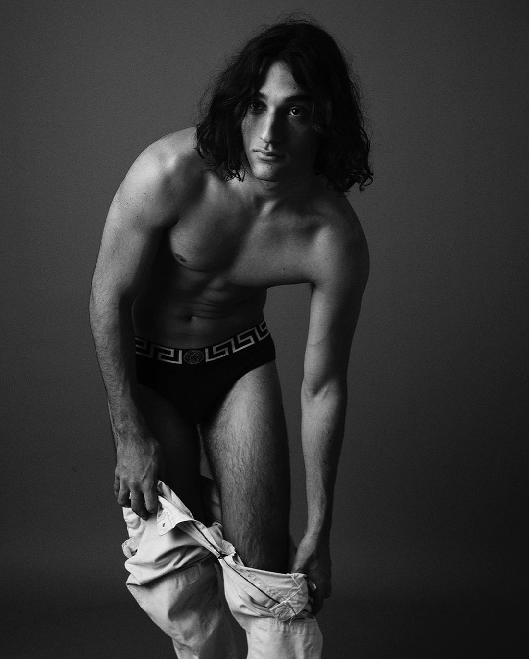 Black and white studio shot of a shirtless adult male with long dark hair pulling down light-colored trousers, staring directly at the camera.