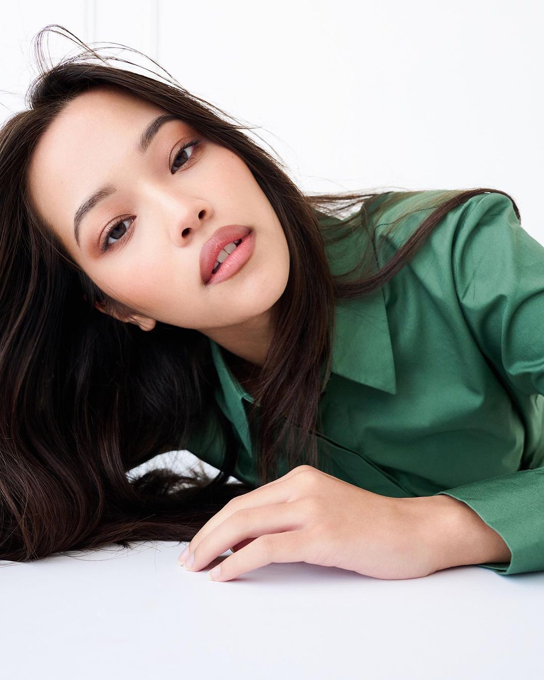 Portrait of a young Asian woman with long dark hair, wearing a green button-down shirt, lying on a white surface in a bright indoor space.