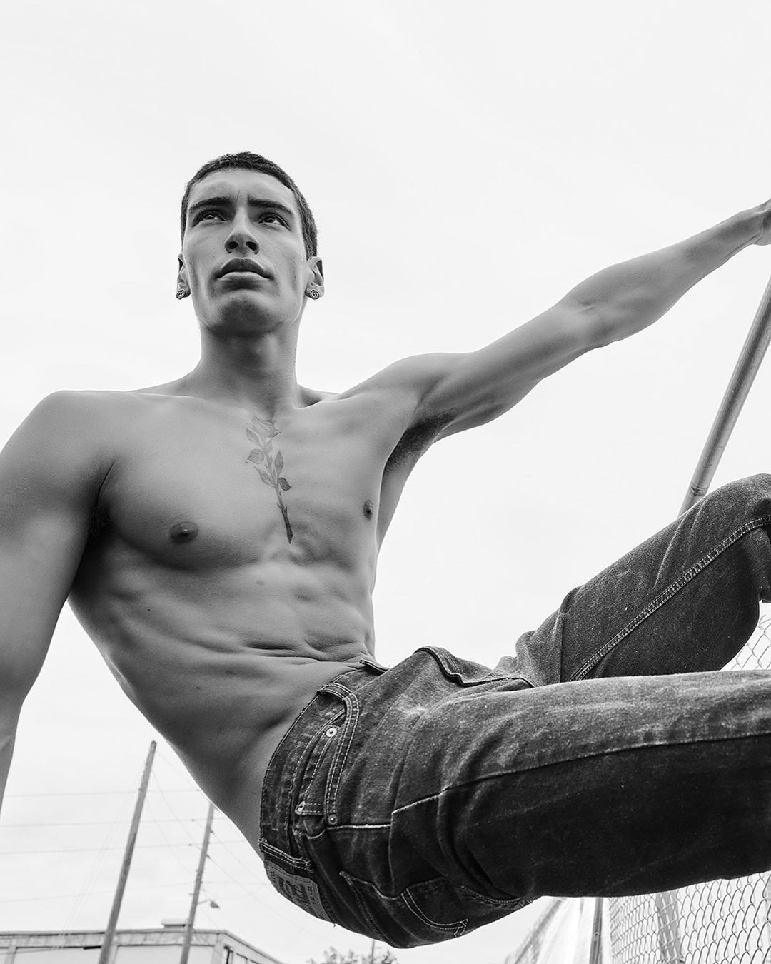 Black and white portrait of a shirtless adult male with visible abs and tattoos, posing outdoors against a bright sky near a chain-link fence.
