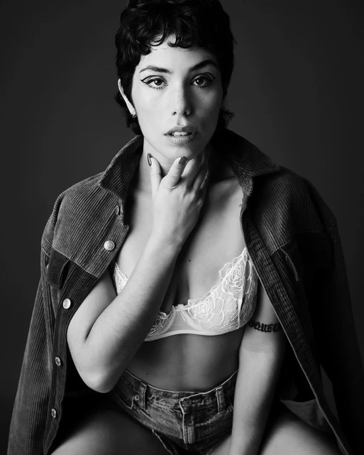 Black and white portrait of a woman with short dark hair wearing a bra, denim shorts, and jacket, seated against a grey background.