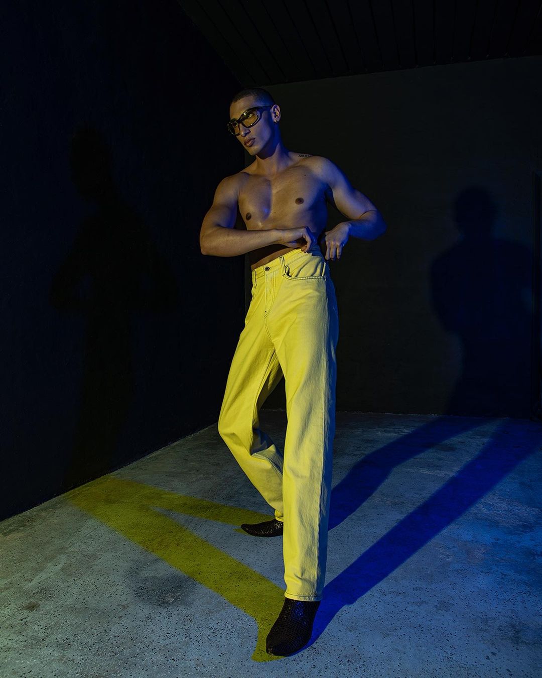 Shirtless man with sunglasses and yellow pants poses indoors with a blue light accenting the concrete floor and dark background.