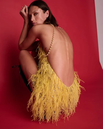 Model crouching in a backless yellow feather dress against a red backdrop, photographed by Juan Algarin