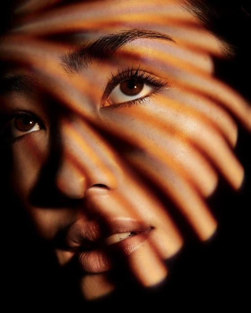 Close-up portrait of an Asian woman's face with dramatic lighting created by orange horizontal blinds casting shadows.