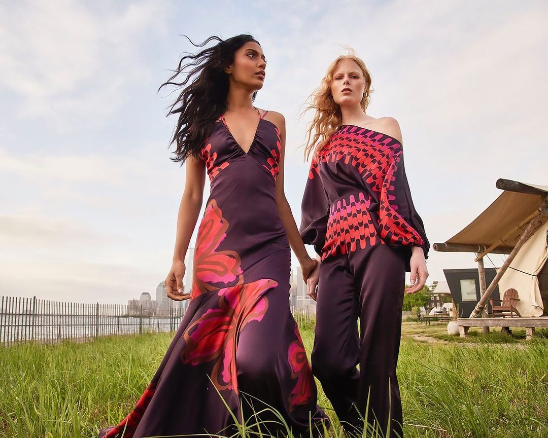 Two women in patterned dresses pose outdoors near a tent and cityscape. They hold hands and look away from the camera.