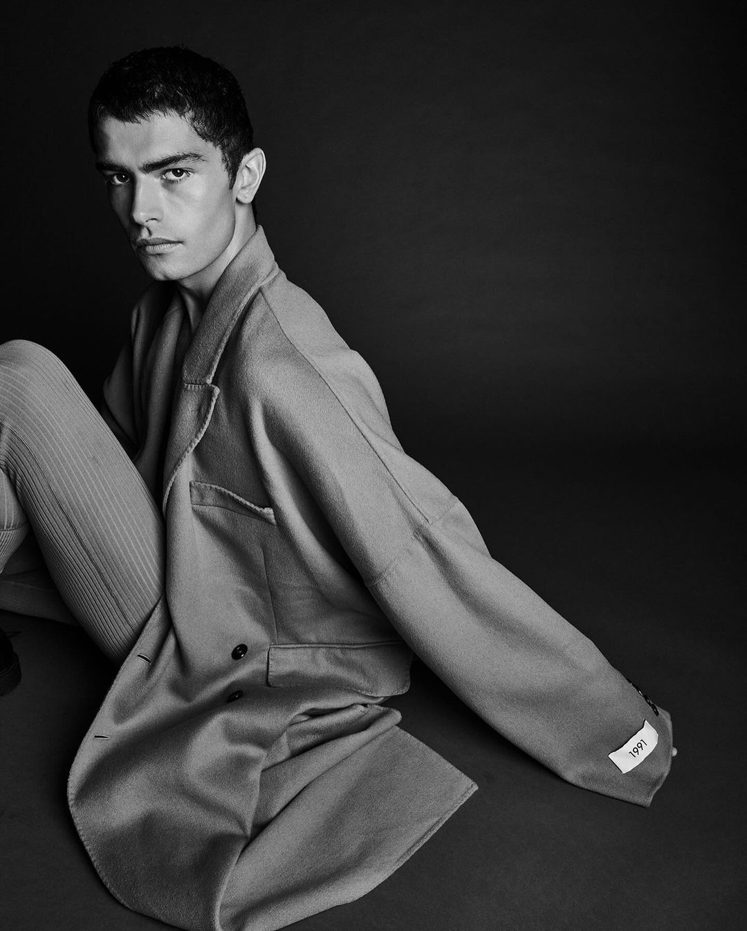 Monochrome portrait of a young male model wearing an oversized coat against a dark background, photographed by Juan Algarin.