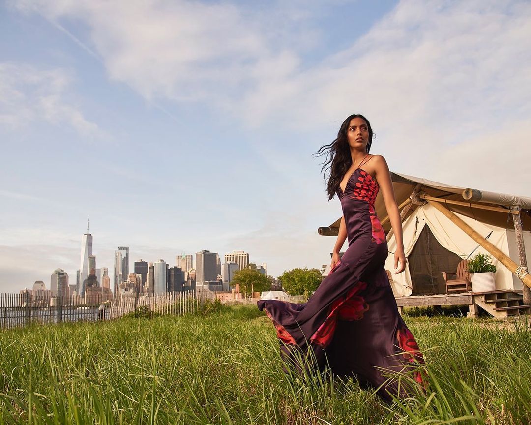 Woman in floral dress stands in grassy field with New York City skyline and tent in background on a sunny day.