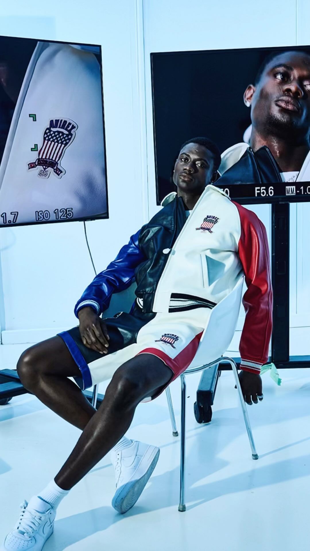 A Black man poses in a color-blocked jacket and shorts while sitting on a white stool in an indoor studio setting with monitors.