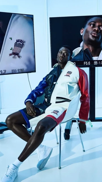 A Black man poses in a color-blocked jacket and shorts while sitting on a white stool in an indoor studio setting with monitors.
