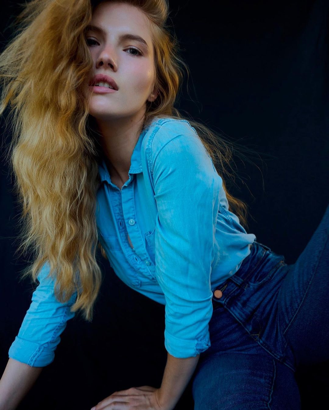 Portrait of a fair-skinned woman with long, wavy blonde hair wearing a light-blue denim shirt and blue jeans against a dark background.