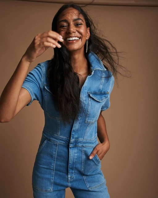 Smiling woman with dark hair poses indoors wearing a blue denim jumpsuit against a brown background in a fashion shot.