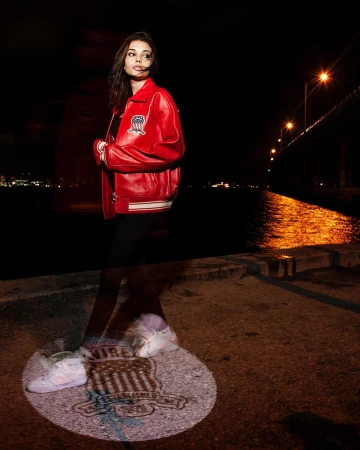 A woman in a red jacket poses at night with a bridge and harbor in the background and a logo projected on the ground.