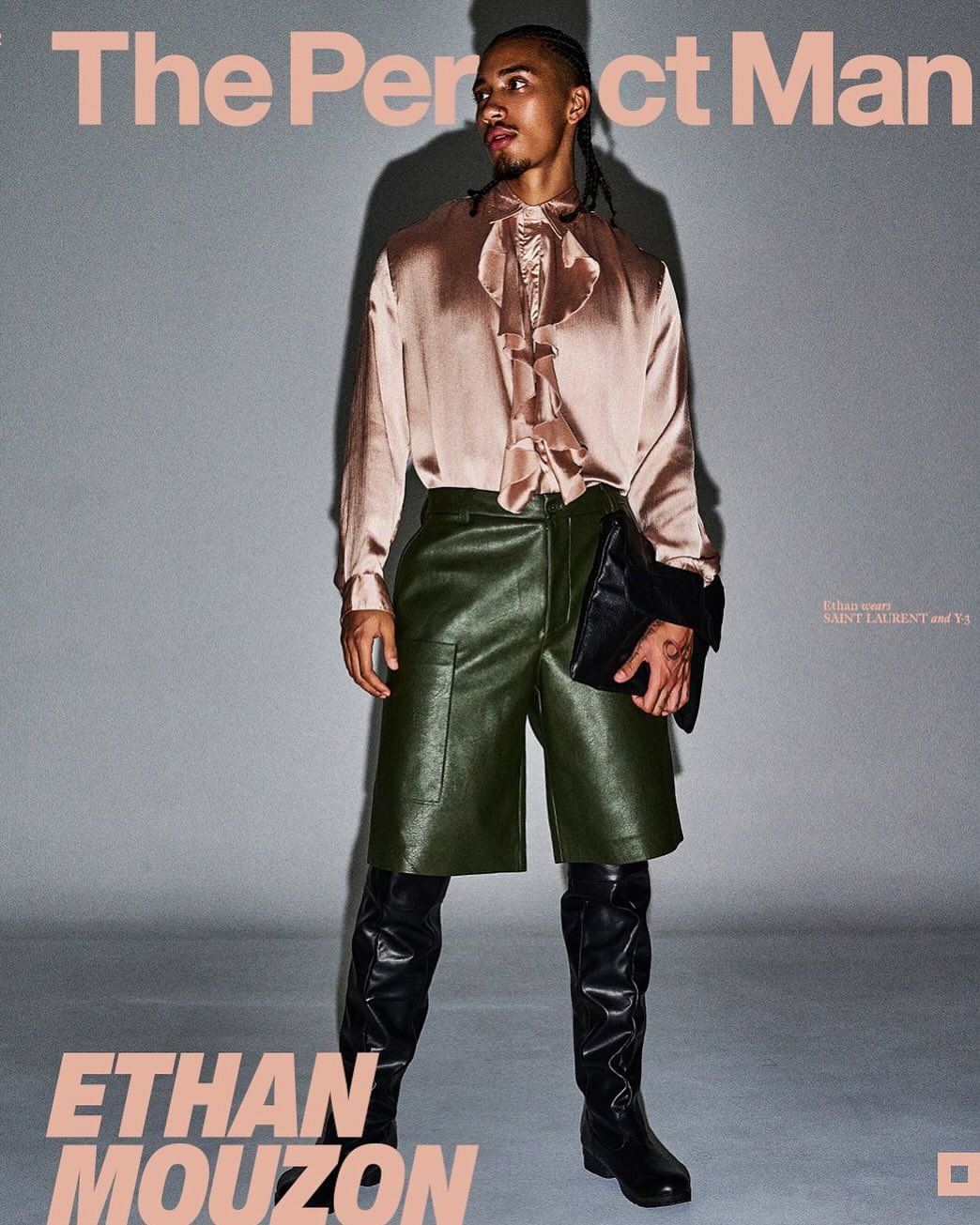 An adult male model wearing a pink ruffled satin shirt, green leather shorts, and high black leather boots poses in a fashion shot.