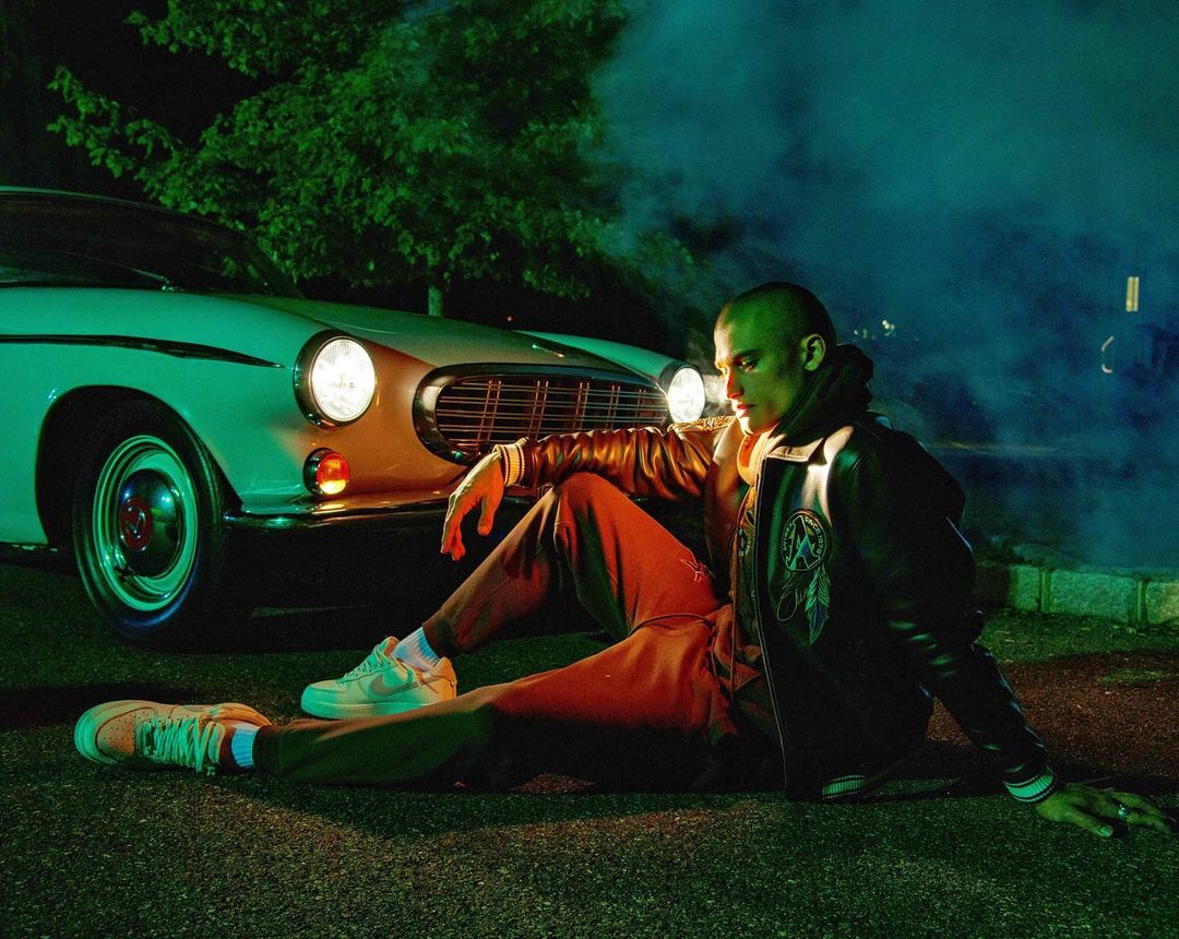 Stylish man in leather jacket sitting on asphalt near vintage car at night with green tree background, captured in medium shot.